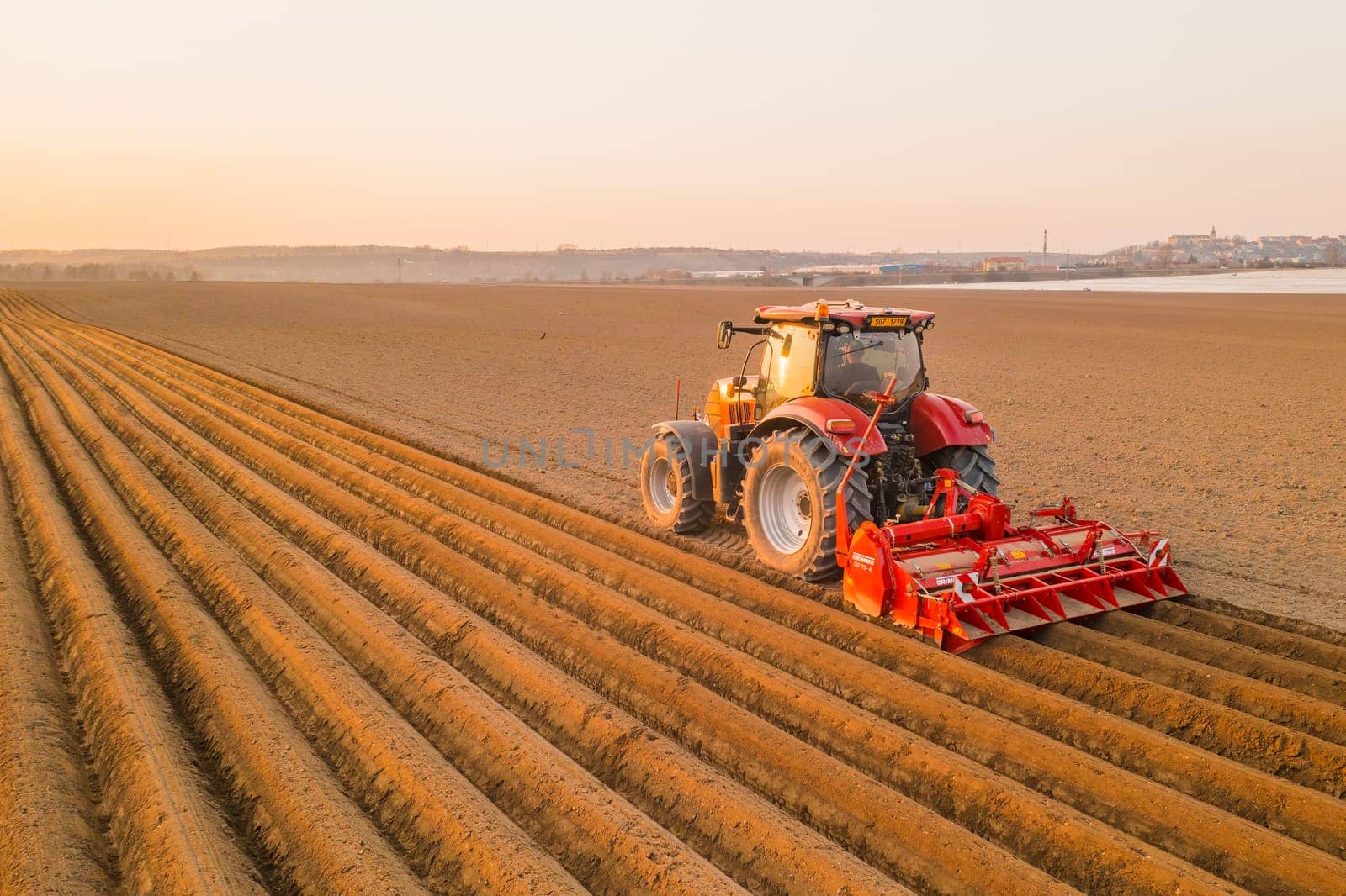 PRAGUE , CZECH REPUBLIC - MARCH 18 2022: Heavy tractor uses plow to make beds for planting potatoes in agricultural field. Powerful machine operates at farm on sunny day aerial view