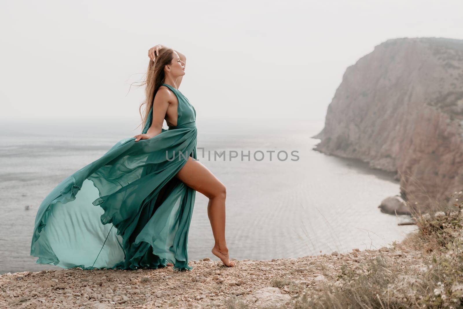 Woman travel portrait. Happy woman with long hair looking at camera and smiling. Close up portrait cute woman in a mint long dress posing on a volcanic rock high above the sea by panophotograph