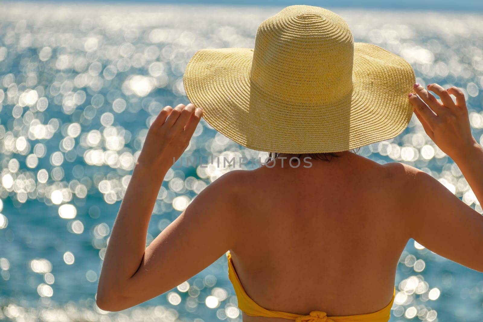 Young woman in a yellow bikini holding on to her hat and looking at the sea.