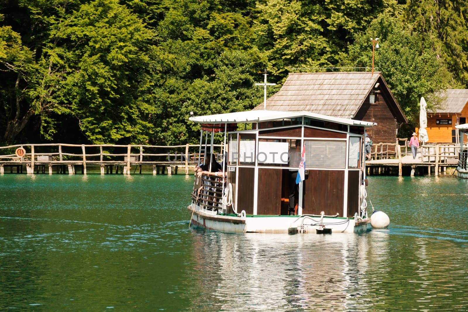 Excursion ship with tourists sails to pier with small country houses against green forest. Touristic attraction in natural park reserve on Plitvice lakes