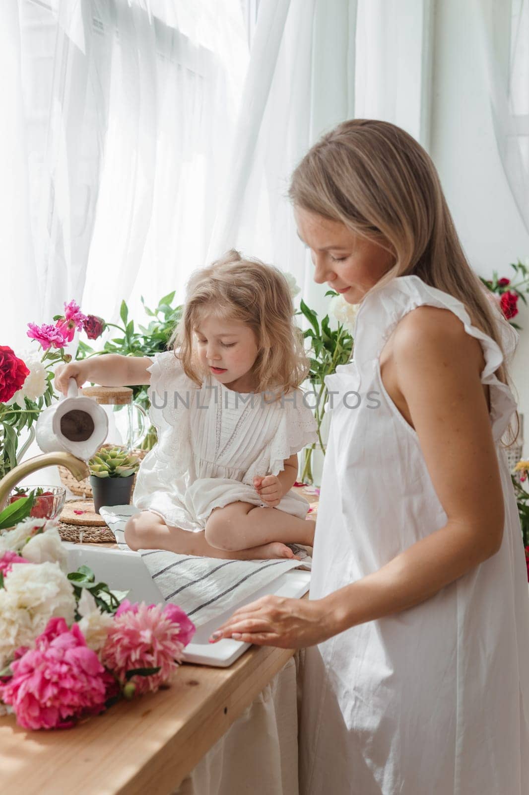 A little blonde girl with her mom on a kitchen countertop decorated with peonies. The concept of the relationship between mother and daughter. Spring atmosphere