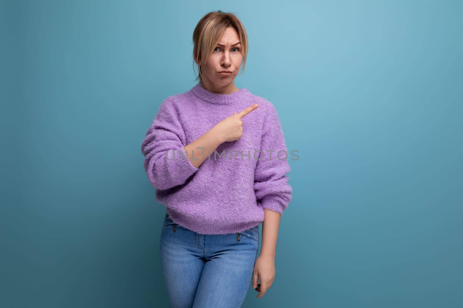 indignant blond young woman in a purple hoodie pointing to the side in surprise on a blue background with copy space.