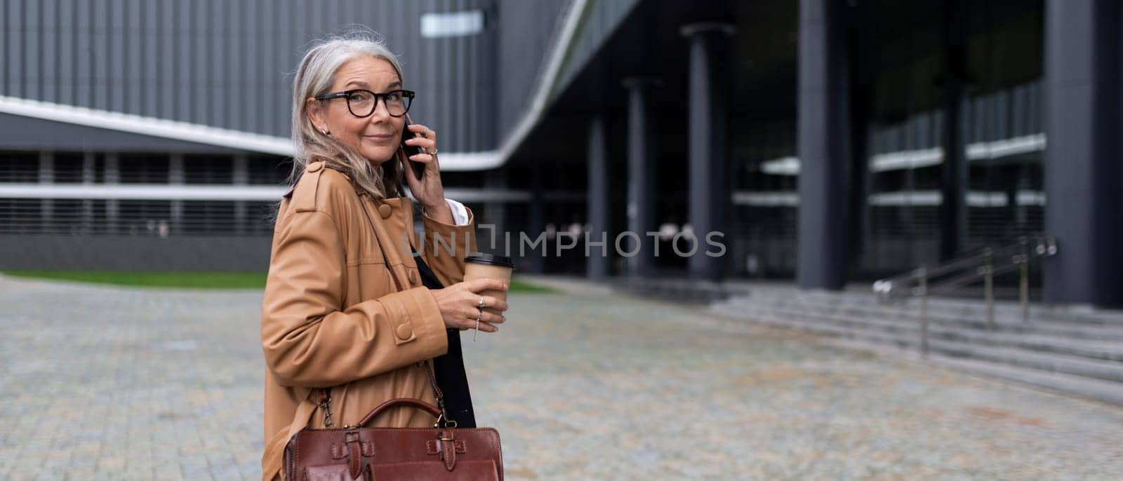 adult successful business woman walks into an office building talking on a mobile phone with a cup of coffee in her hands, caucasian Middle aged older businesswoman at work Concept by TRMK