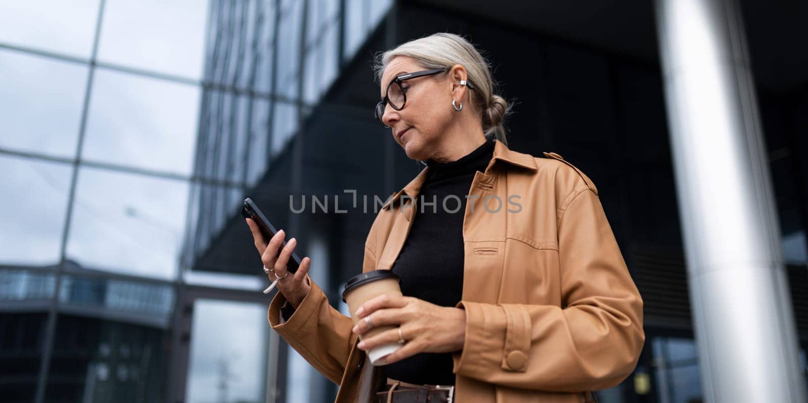 stylish elderly woman with a mobile phone and a glass of coffee on the background of a modern business center, career growth concept by TRMK