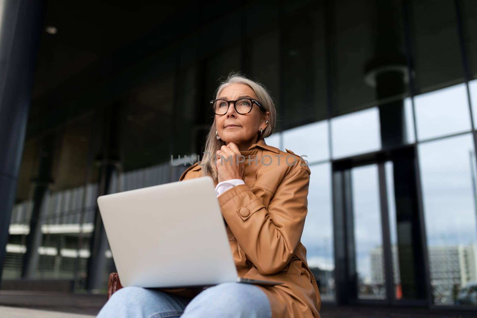 a businesswoman sits with a laptop on her knees on the steps at the entrance to an office building.