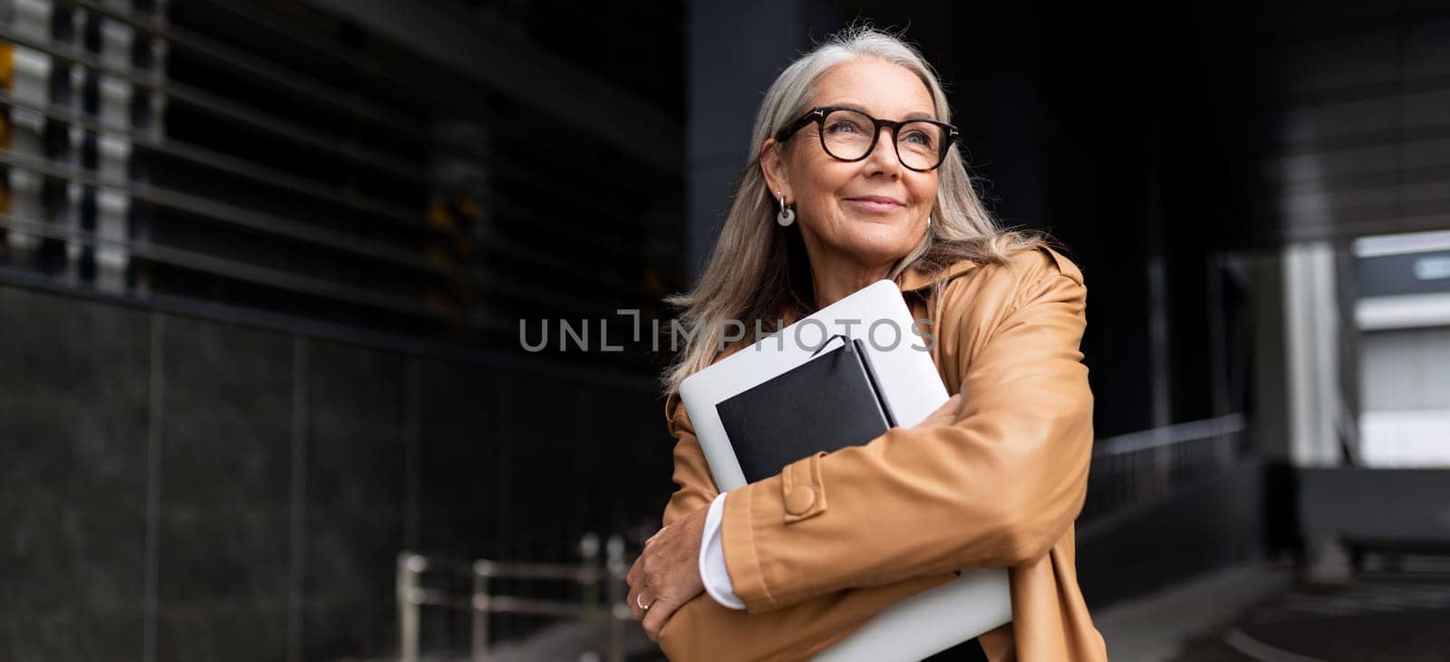 portrait of an elderly businesswoman with a laptop in glasses outside the office, strong and independent woman concept by TRMK