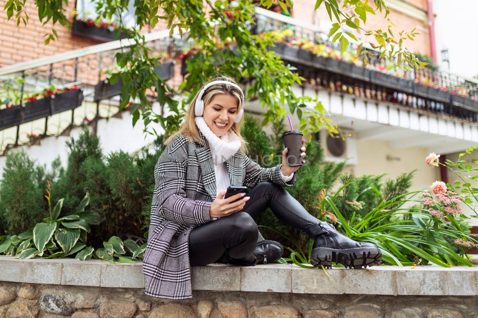 caucasian woman in autumn coat listens to music on headphones and looks at mobile phone outside by TRMK