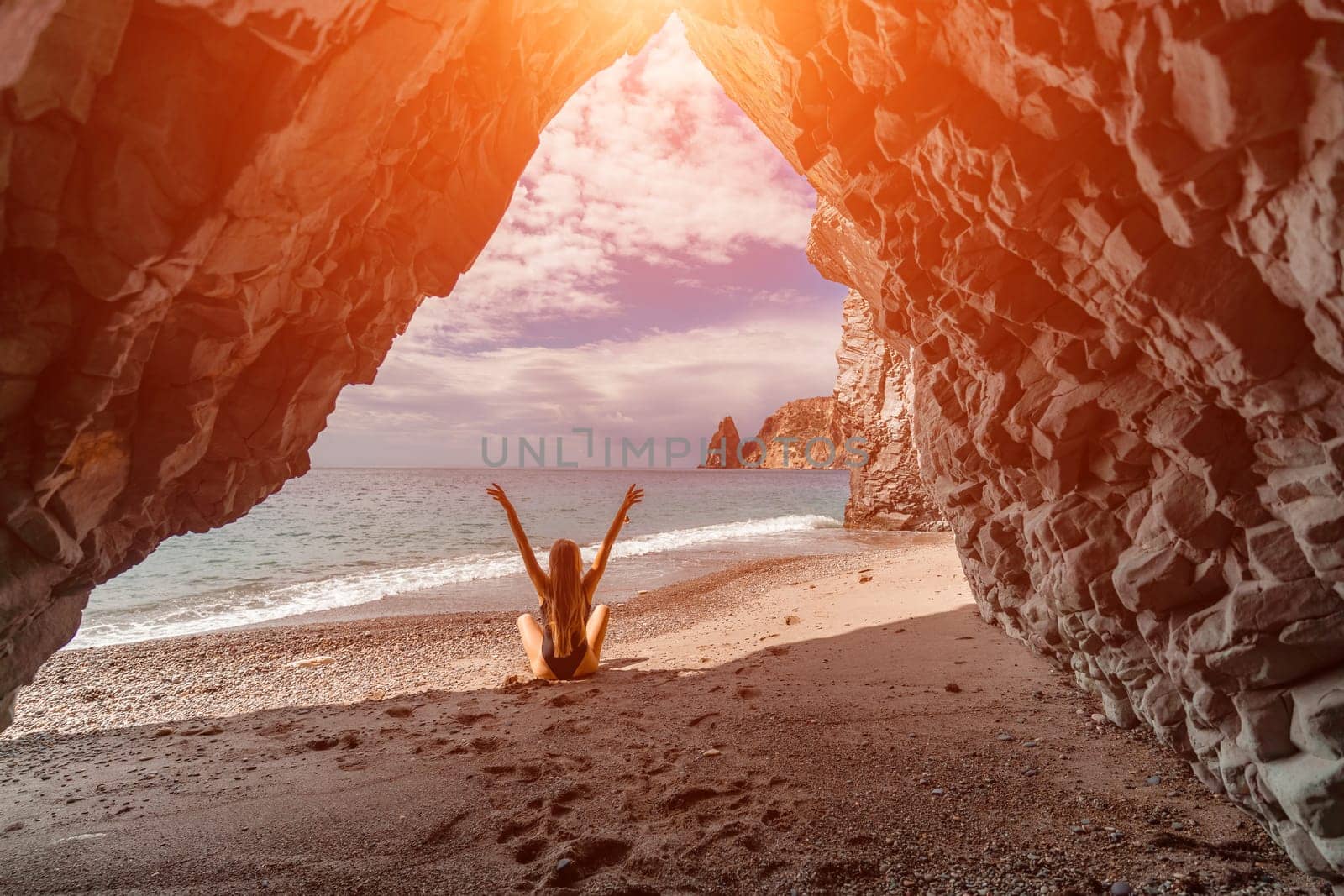 View of a woman in a black swimsuit from a sea cave Attractive woman enjoying the sea air sits on the beach and looks at the sea. by Matiunina