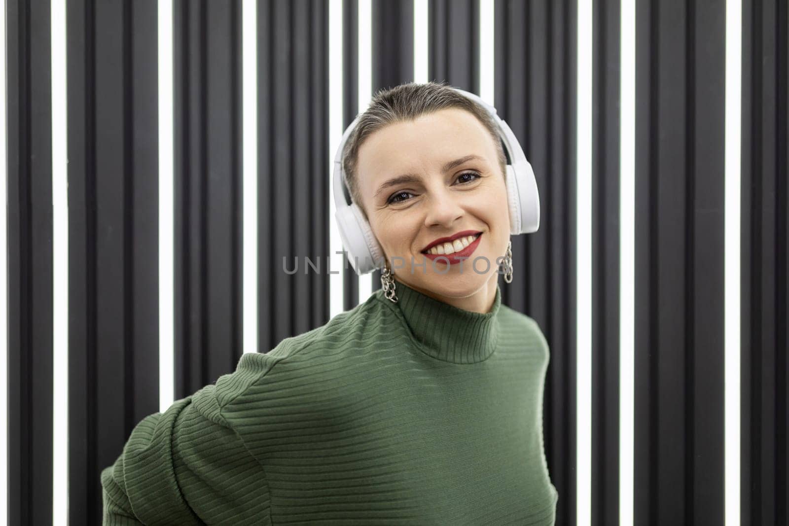 a cheerful mature woman with a short haircut in headphones with a wide smile on the background of a white-black wall looks at the camera by TRMK