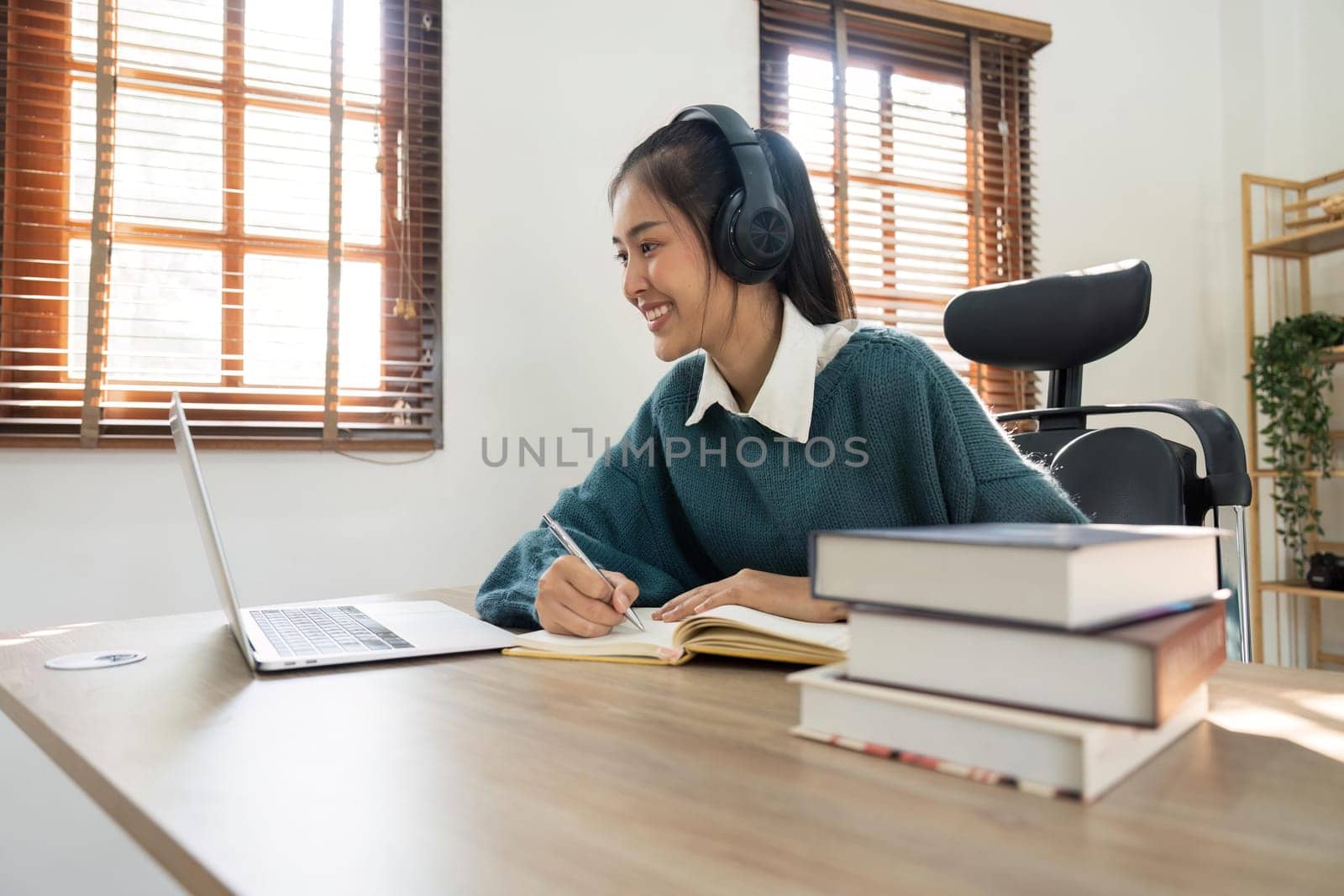 Smiling girl student wear wireless headphone study online with teacher, happy young woman learn language listen lecture watch webinar write notes look at laptop, distant education.