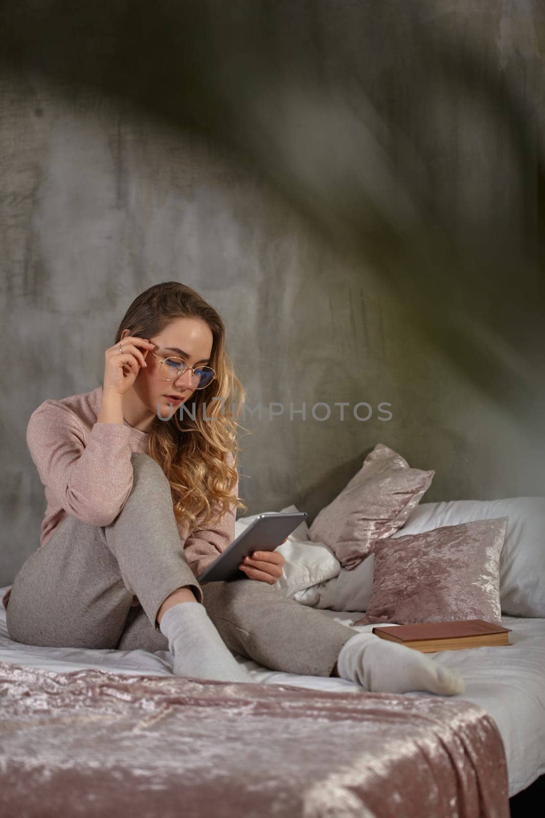 Gorgeous blonde lady in glasses, casual clothing. Sitting on bed, working with a tablet in bedroom at home. Student, blogger, document studying. Interior with gray wall, pink blanket and pillows