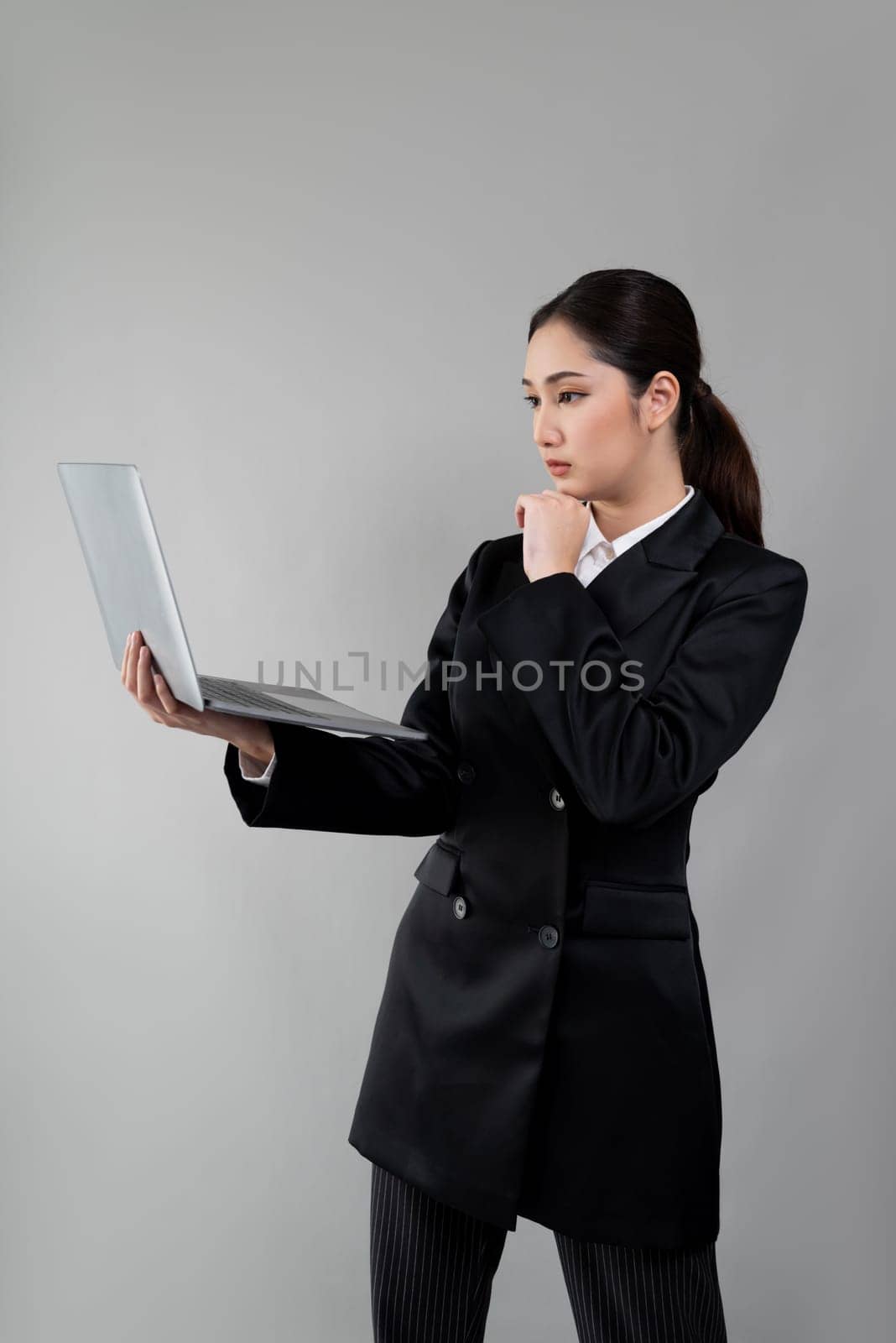 Confident young businesswoman stands on isolated background, working on laptop posing in formal black suit. Office lady or manager with smart and professional appearance. Enthusiastic