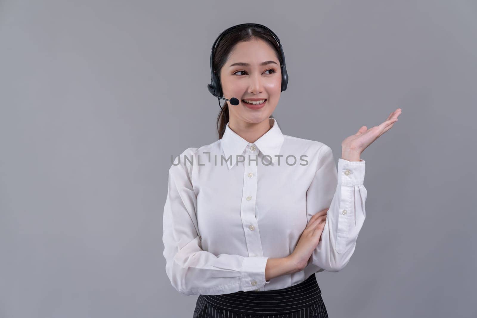 Asian female call center operator with smile face advertises job opportunity, wearing a formal suit and headset holding hand gesture for product on customizable isolated background. Enthusiastic