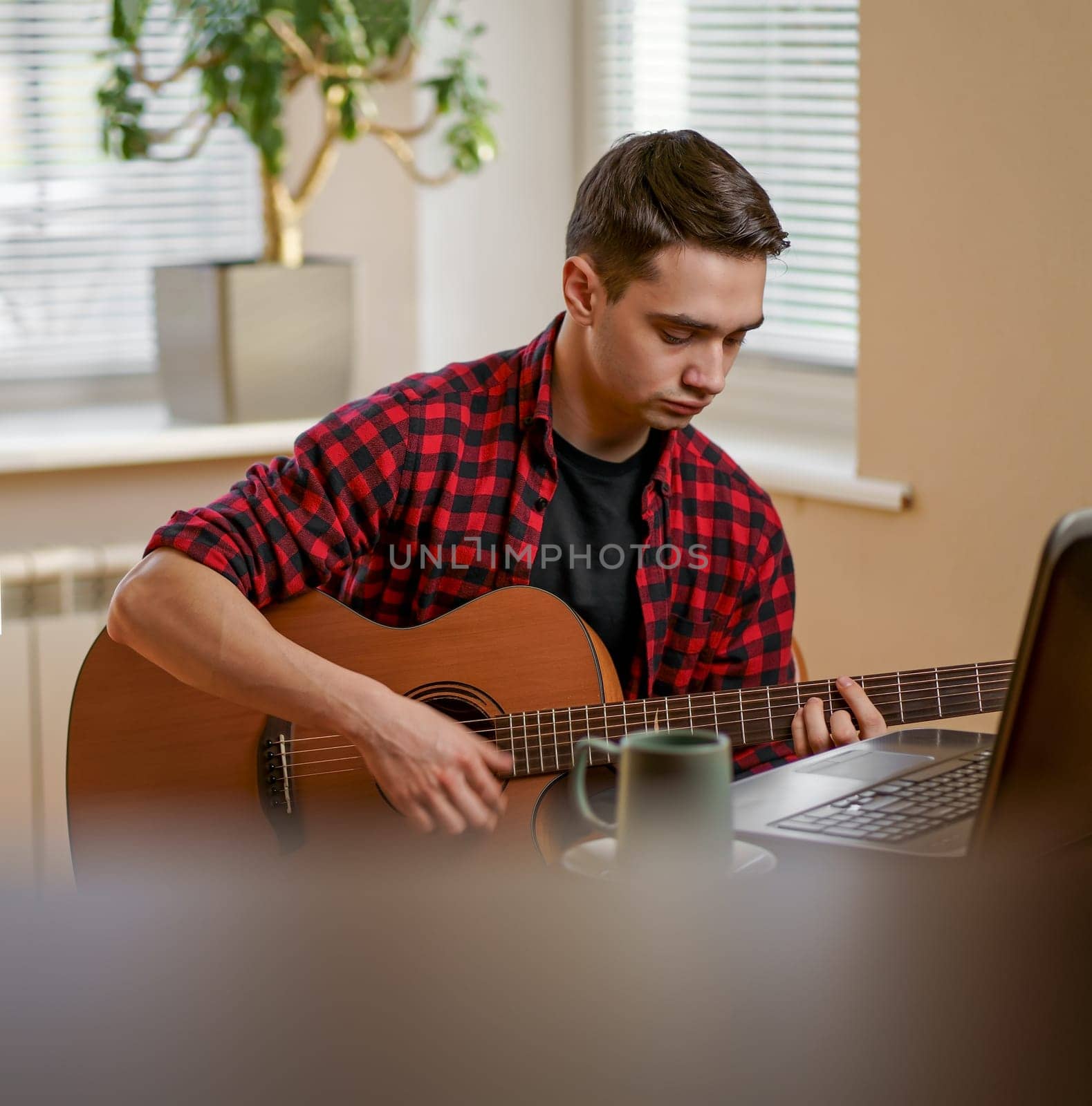 Relaxed young man playing guitar at home and using laptop, drinking tea by aprilphoto