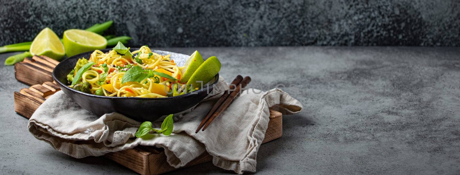 Asian vegetarian noodles with vegetables and lime in black rustic ceramic bowl, wooden chopsticks on cutting board angle view on stone background. Cooking noodles by its_al_dente