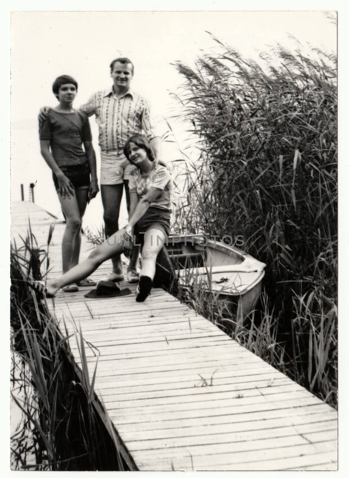 GREAT LAKES OF MECKLENBURG, GERMANY - AUGUST 1977 - Vintage photo shows father and adolescent - teenager children pose on the pier. The pier and boat - summer holidays - vacation theme. Circa 1980.