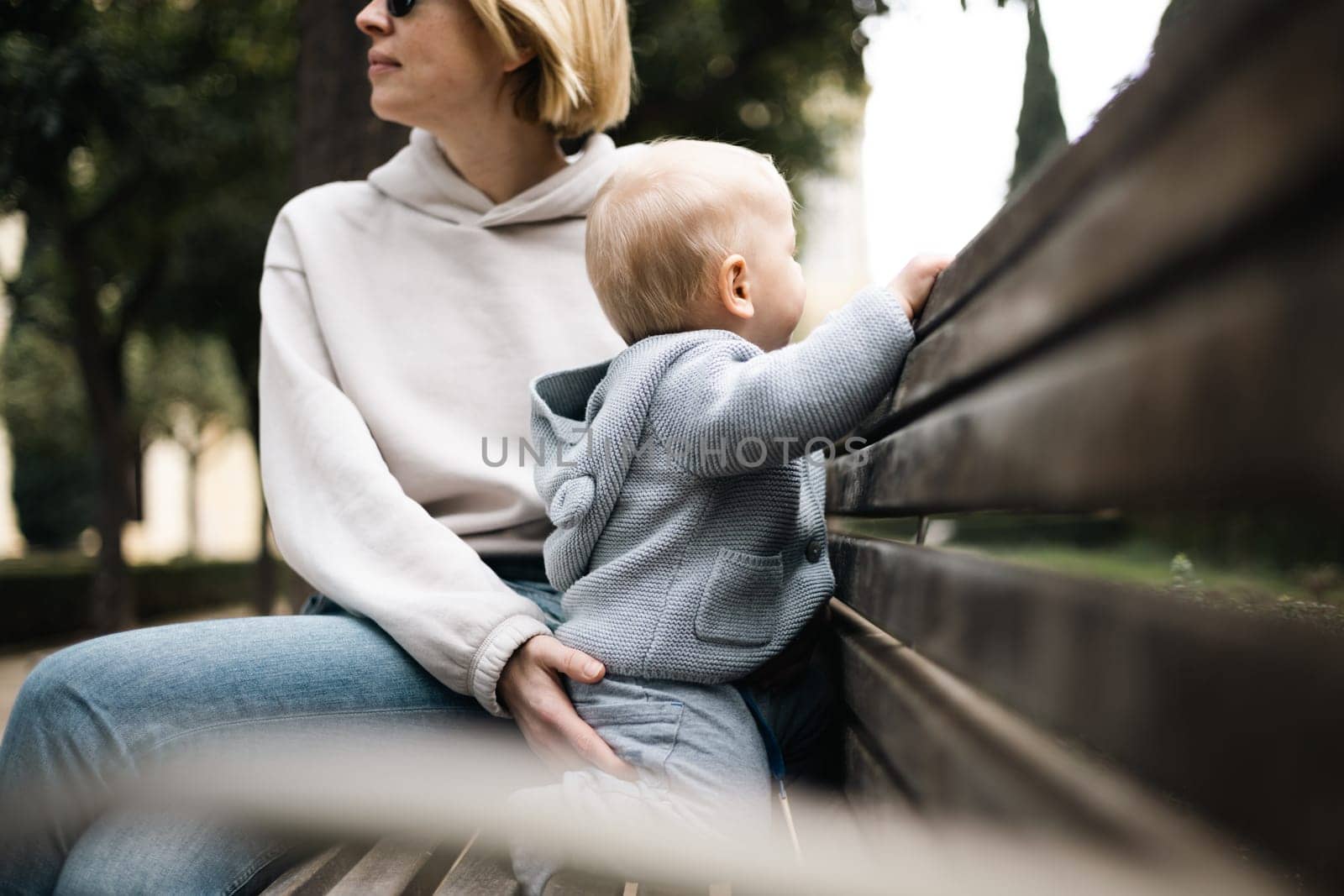 Young mother with her cute infant baby boy child sitting on bench in city park