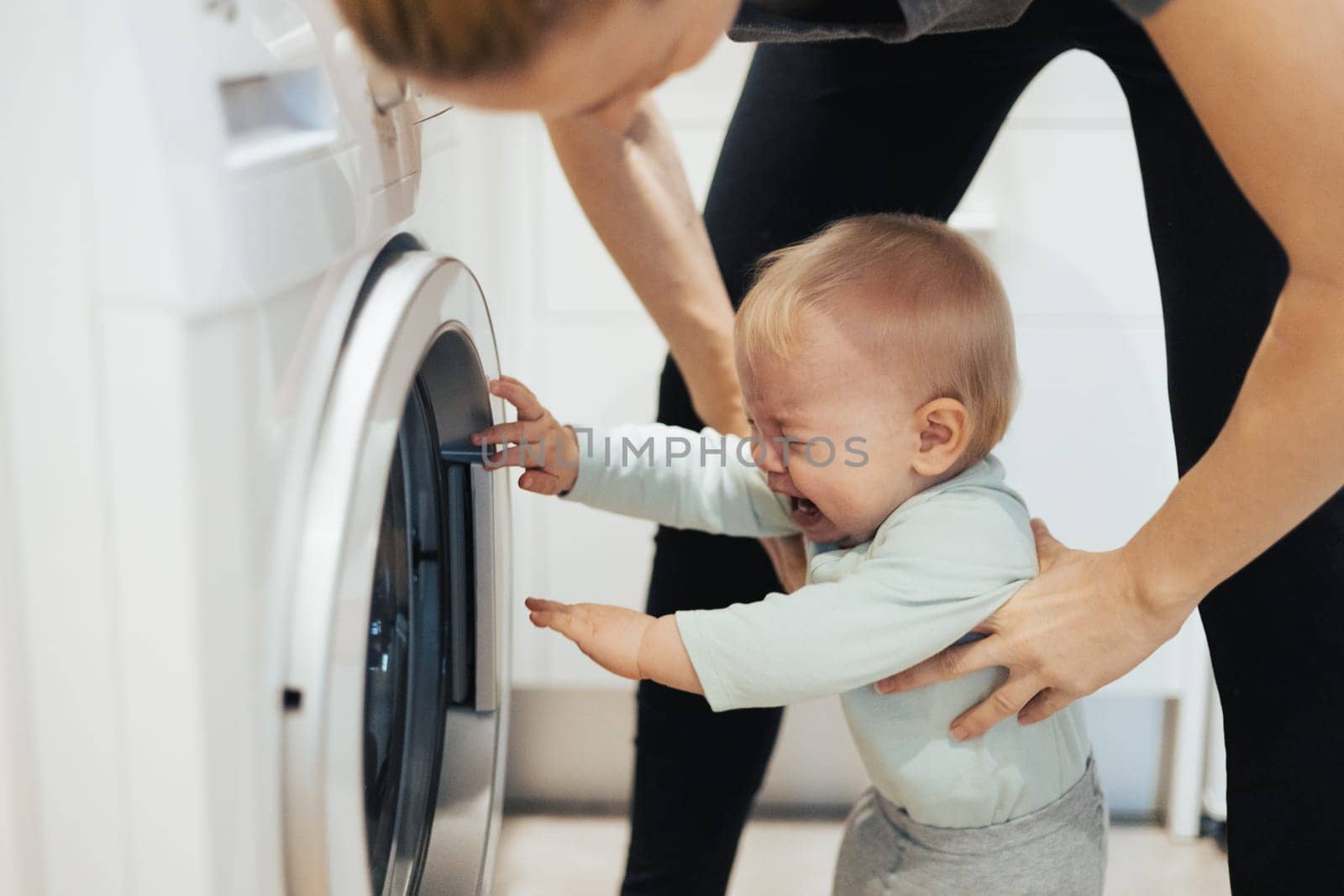 Little infant baby boy child demanding mother's attention while she is multitasking, trying to do some household chores in kitchen at home. Mother on maternity leave