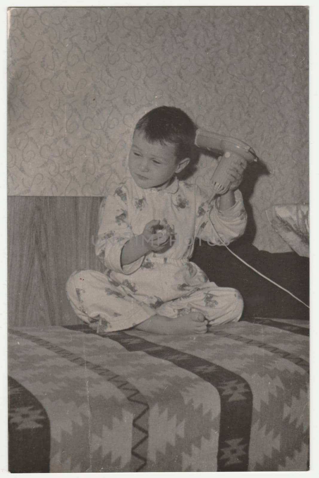 Vintage photo shows a small boy wears pyjamas and he uses hair dryer. Funny photography of a cute boy. Retro black and white photography. Circa 1970. by roman_nerud