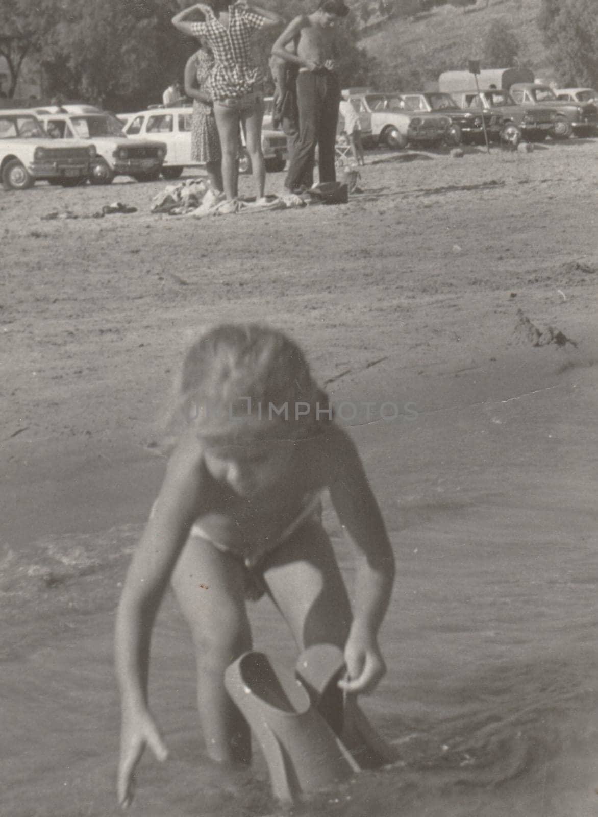 Vintage photo shows a small girl plays in the water with swimming fins. Holidays - vacation theme. Retro black and white photography. Circa 1980. by roman_nerud