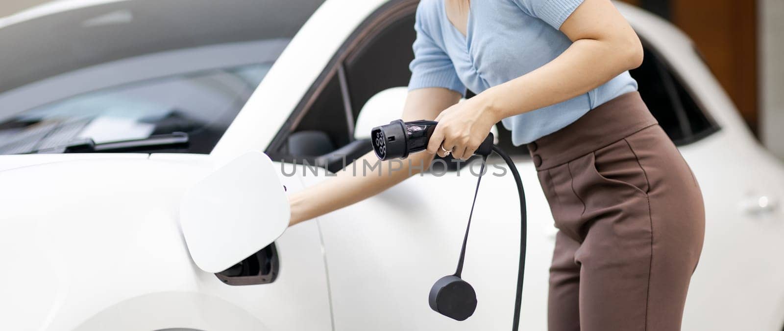 Closeup progressive woman install cable plug to her electric car with home charging station. Concept of the use of electric vehicles in a progressive lifestyle contributes to clean environment.