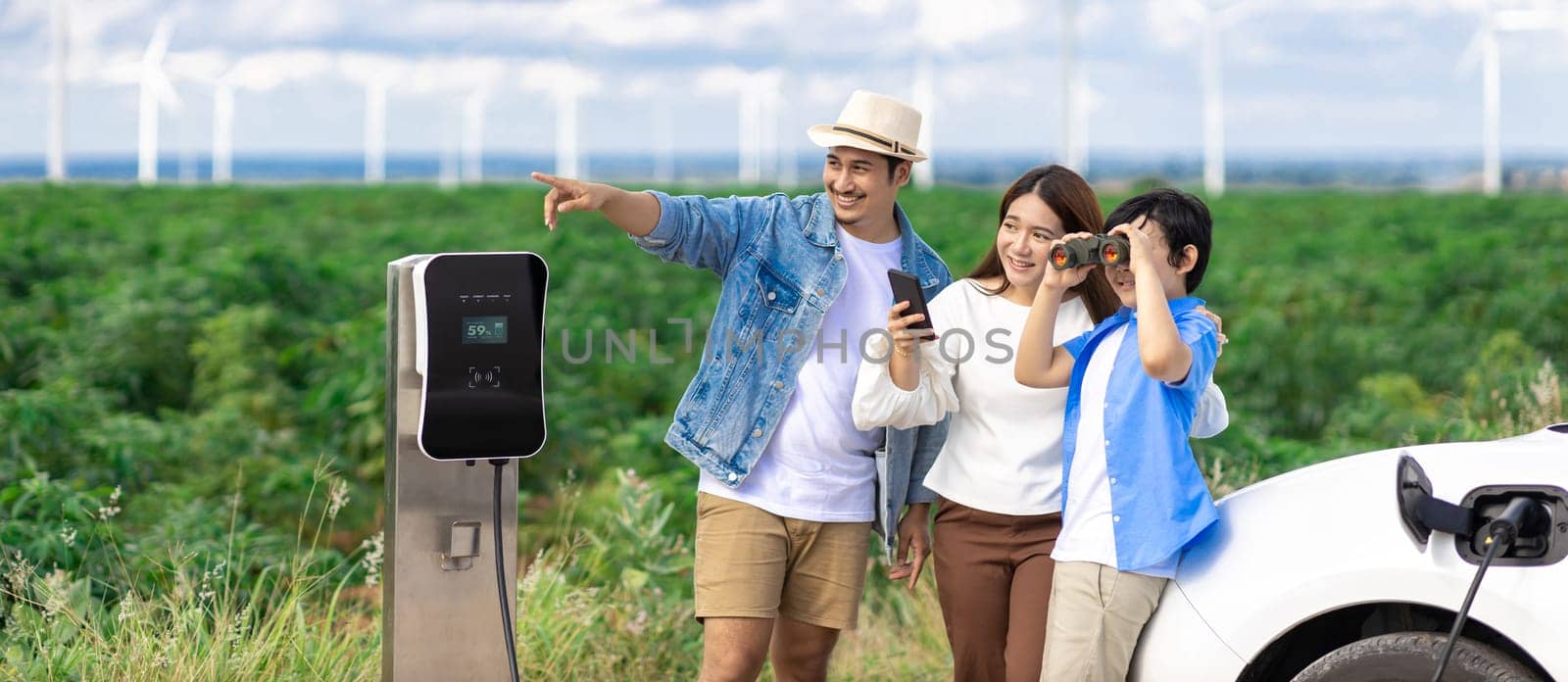 Concept of progressive happy family enjoying their time at wind farm with electric vehicle. Electric vehicle driven by clean renewable energy from wind turbine generator for charging station.