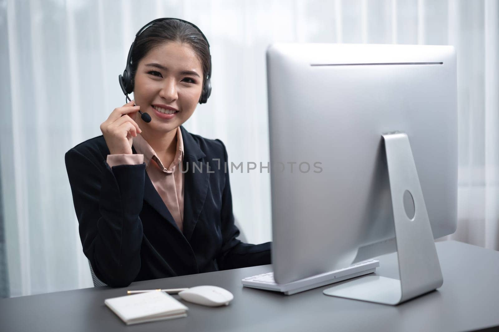 Asian call center with headset and microphone working on her workspace looking at camera. Female operator provide exceptional customer service. Supportive call center agent portrait. Enthusiastic