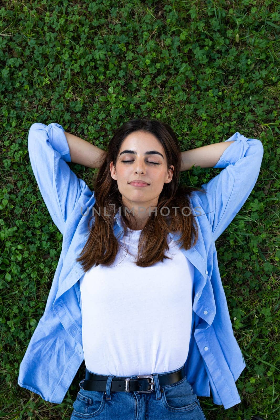 Top view vertical portrait of serene, relaxed young brunette woman lying down on grass with eyes closed. Wellbeing and lifestyle concept.