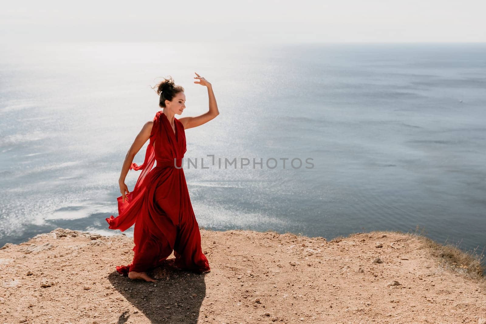 Side view a Young beautiful sensual woman in a red long dress posing on a rock high above the sea during sunrise. Girl on the nature on blue sky background. Fashion photo.