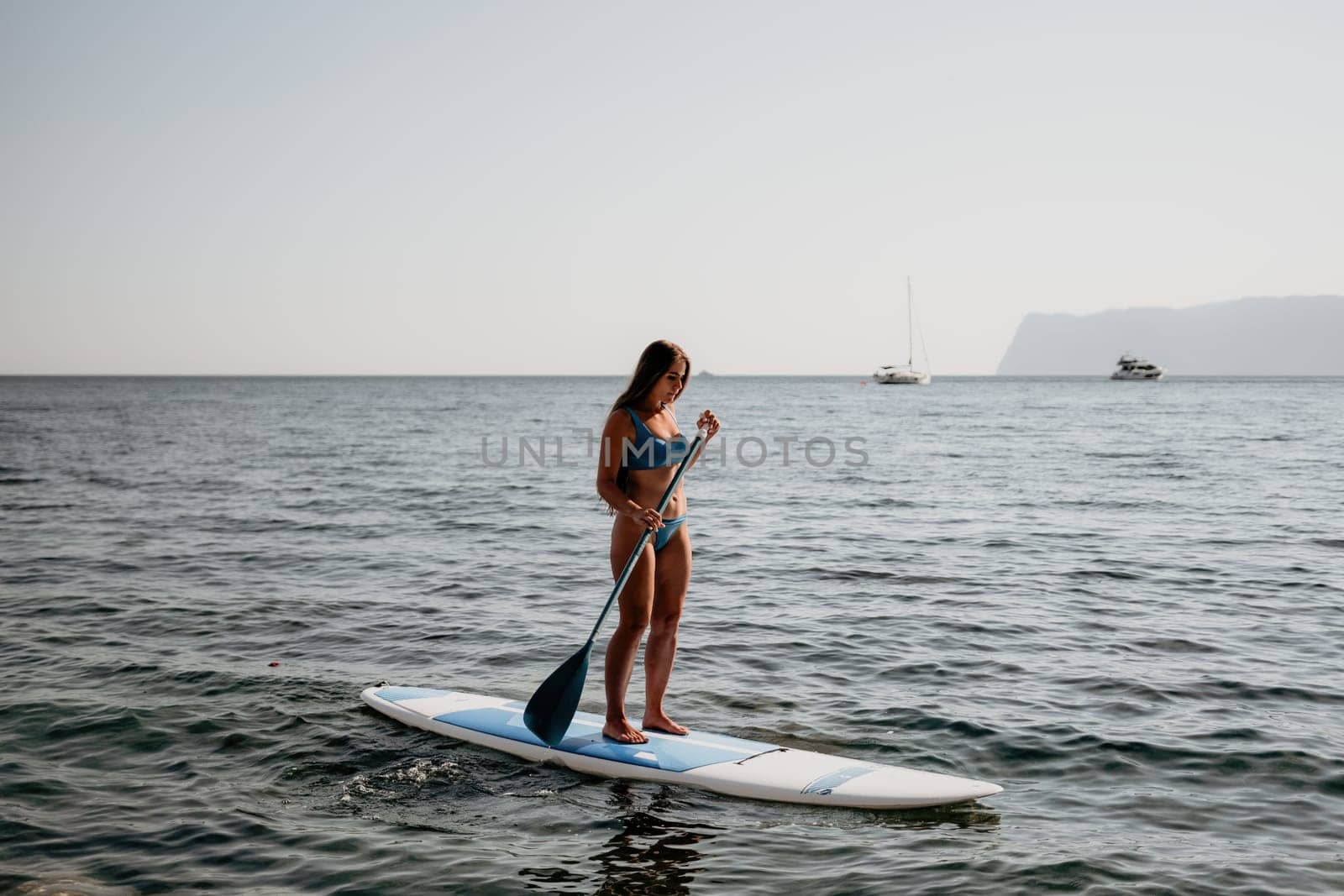 Woman sea sup. Close up portrait of happy young caucasian woman with long hair looking at camera and smiling. Cute woman portrait in a blue bikini posing on sup board in the sea by panophotograph