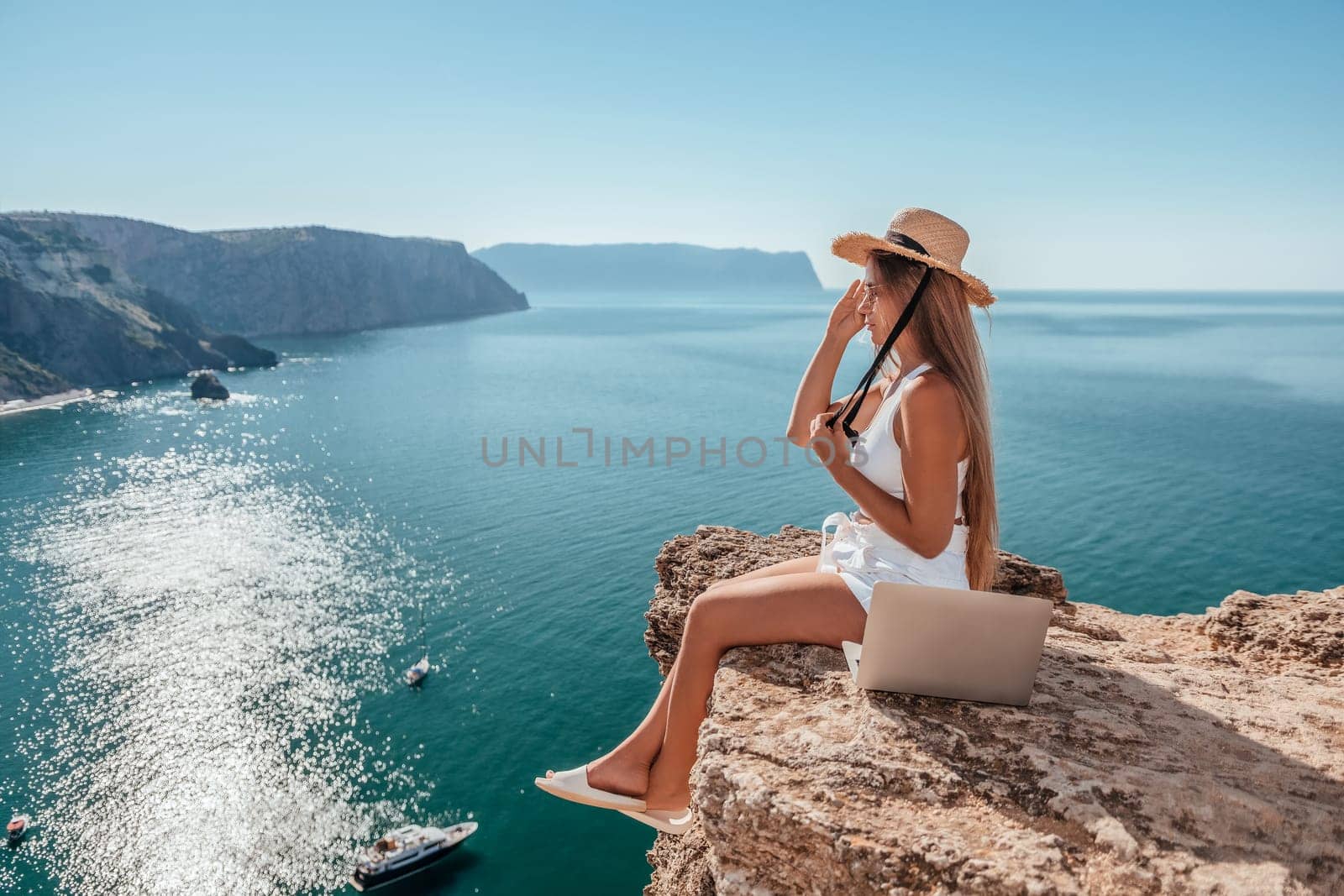 Woman laptop sea. Working remotely on seashore. Happy successful woman female freelancer in straw hat working on laptop by the sea at sunset. Freelance, remote work on vacation by panophotograph