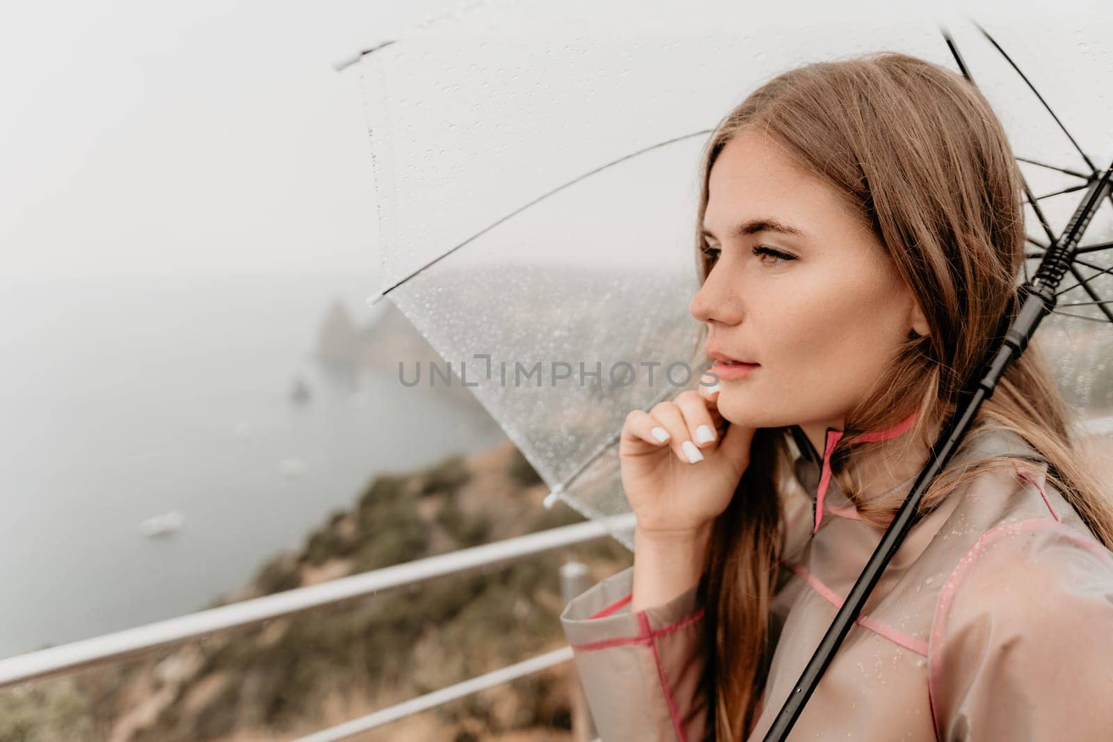 Woman rain park. Happy woman portrait wearing a raincoat with transparent umbrella outdoors on rainy day in park near sea. Girl on the nature on rainy overcast day