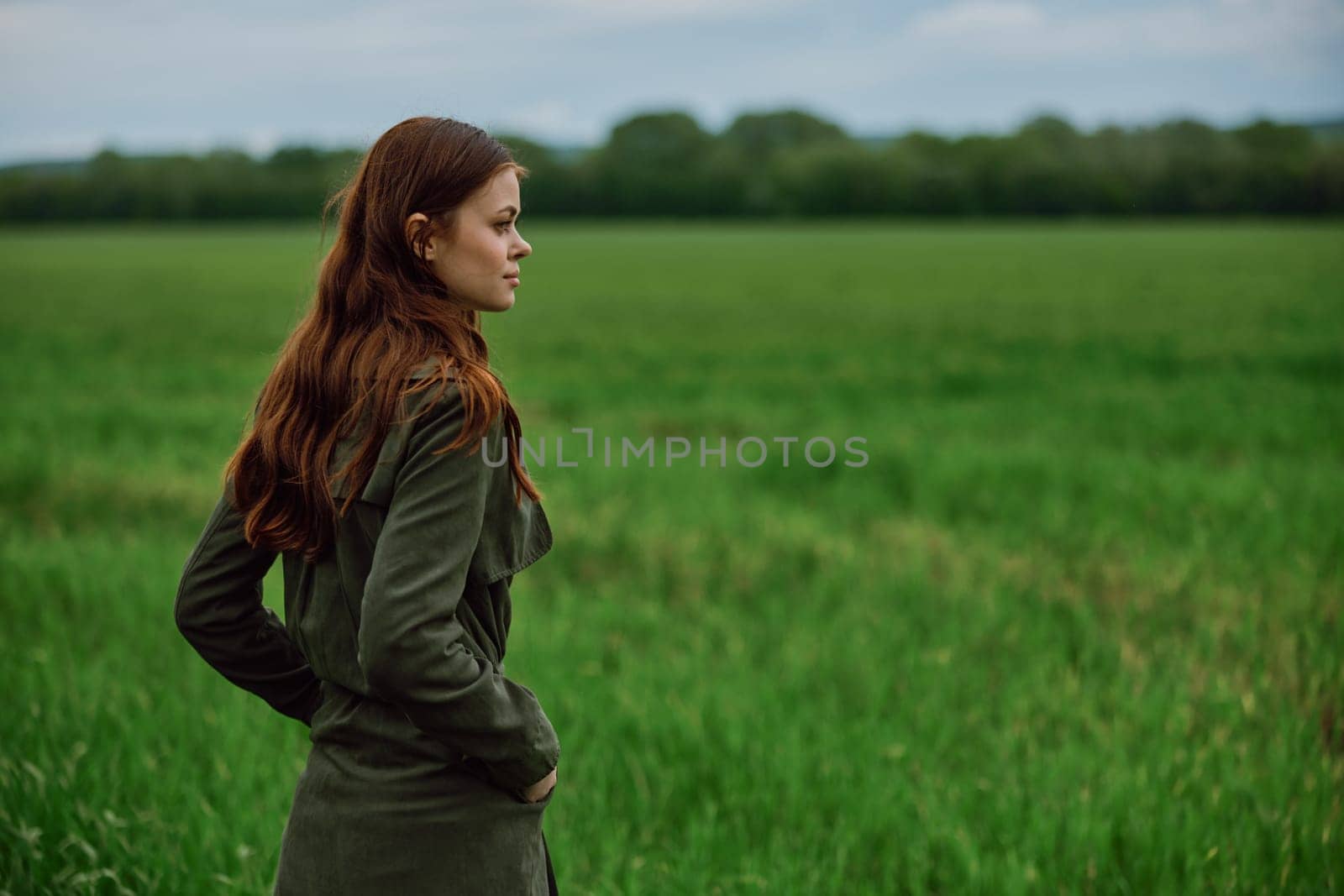 a beautiful, red-haired girl in a raincoat stands in a field in the spring in rainy weather. High quality photo