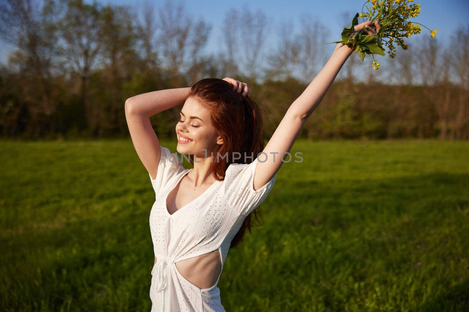 a beautiful, happy woman in a light dress stands in a field raising her hands high holding a bouquet of flowers by Vichizh