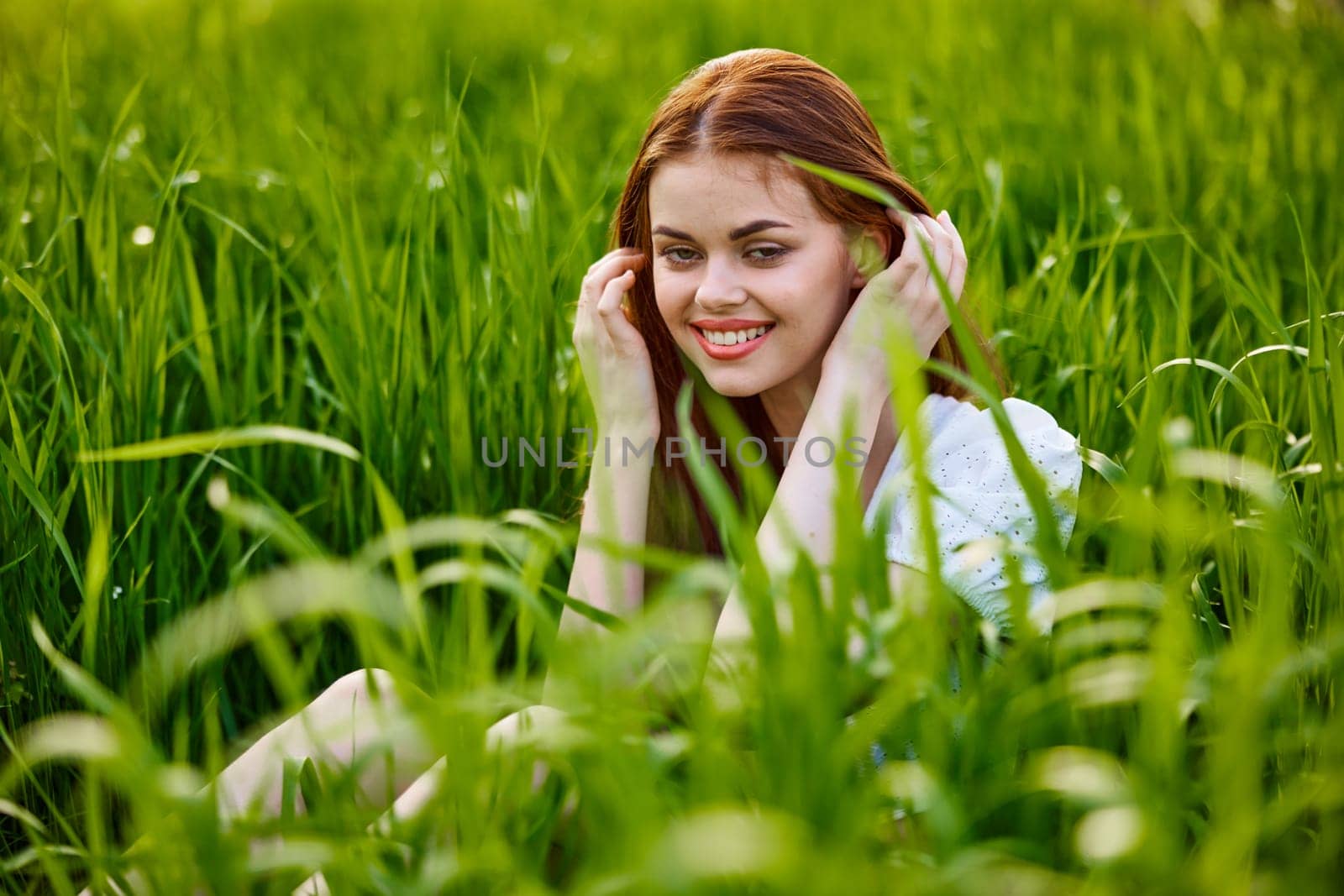 beautiful, elegant woman with red hair sits in tall green grass and smiles by Vichizh