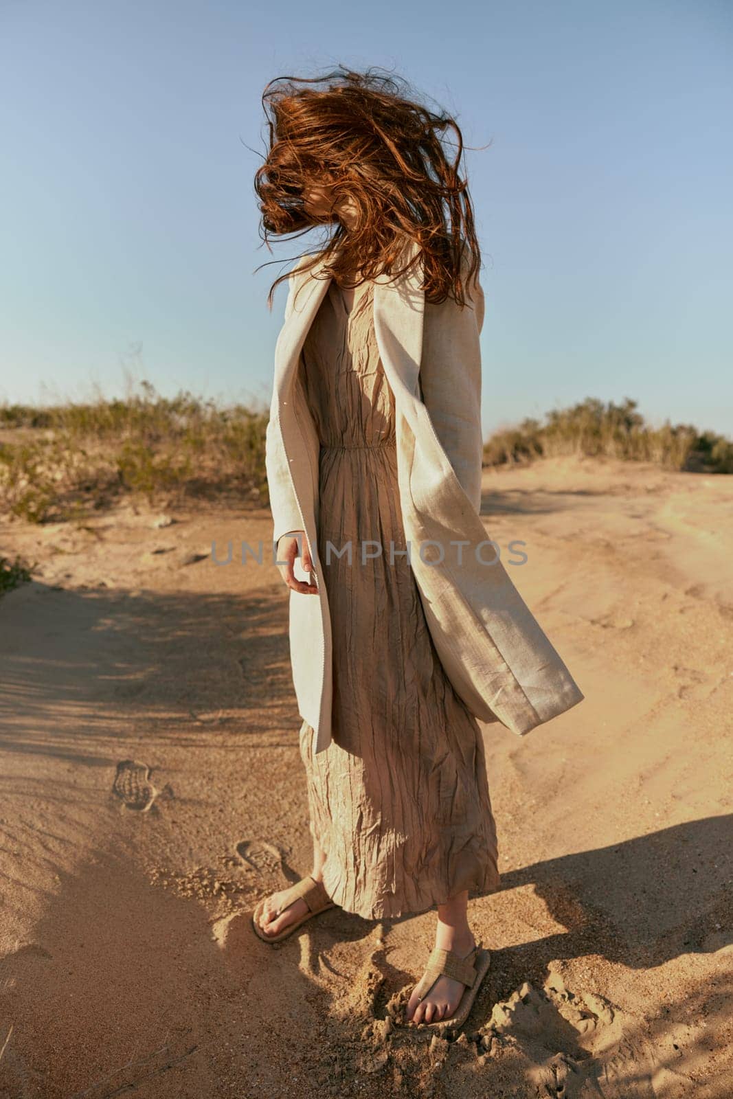 a woman in a light jacket poses on the sand with her face covered by her hair from a strong wind by Vichizh