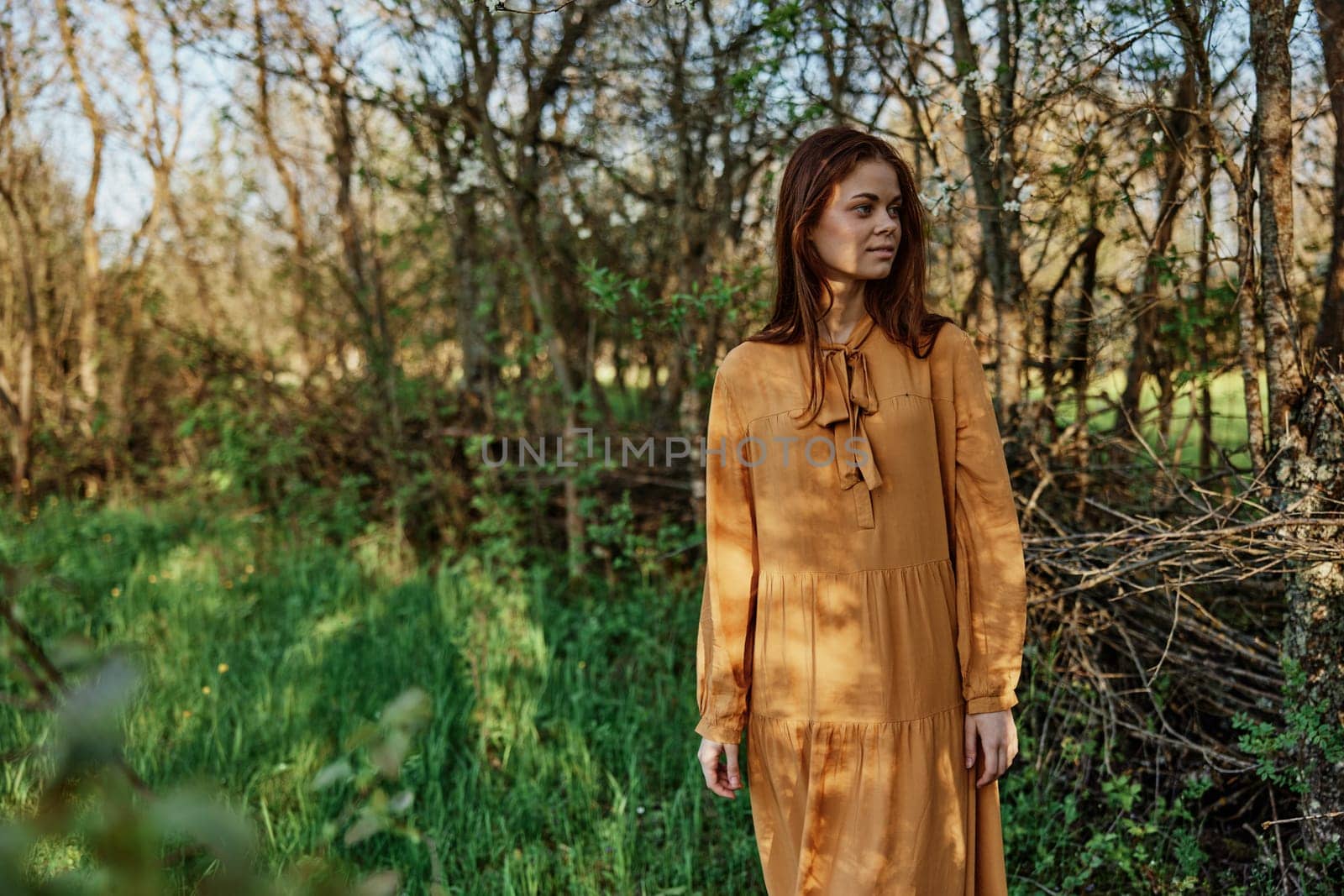 a beautiful, slender woman with long hair walks in the shade near the trees, dressed in a long orange dress, enjoying the weather and the weekend. High quality photo