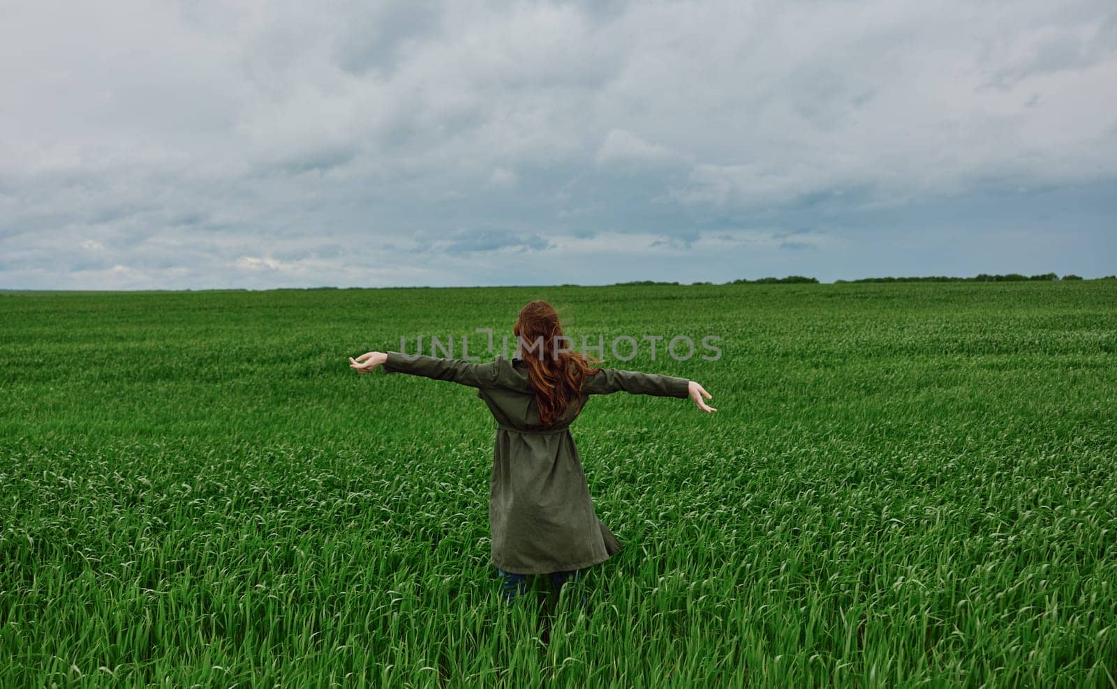 a woman in a long coat stands in a green field on a cloudy day with her hands up to the sky by Vichizh