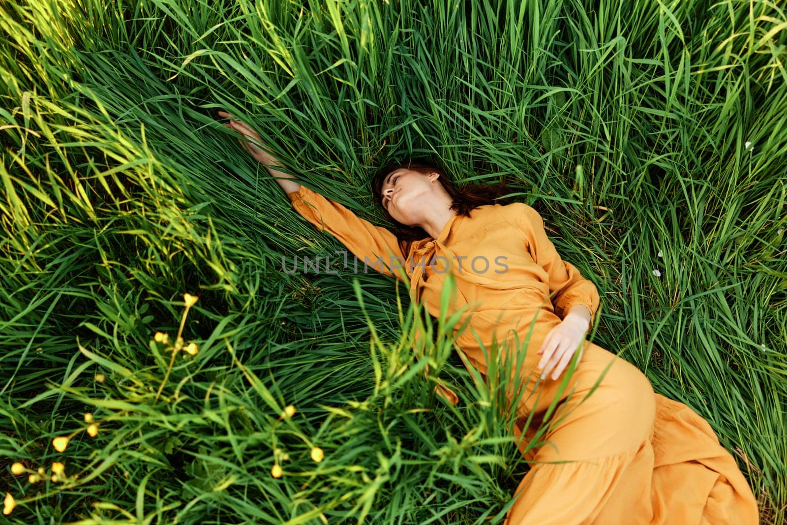 a relaxed woman enjoys summer lying in the tall green grass in a long orange dress stretched out stretching her arm forward by Vichizh