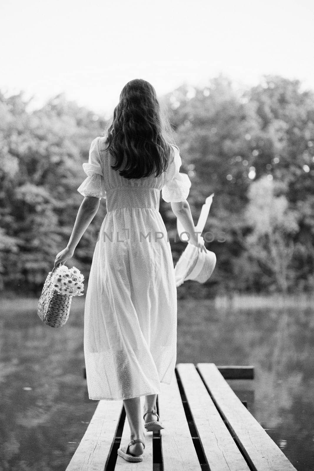 a woman in a long light dress stands on a pier by a lake in the forest with a basket and a wicker hat. monochrome photography. High quality photo