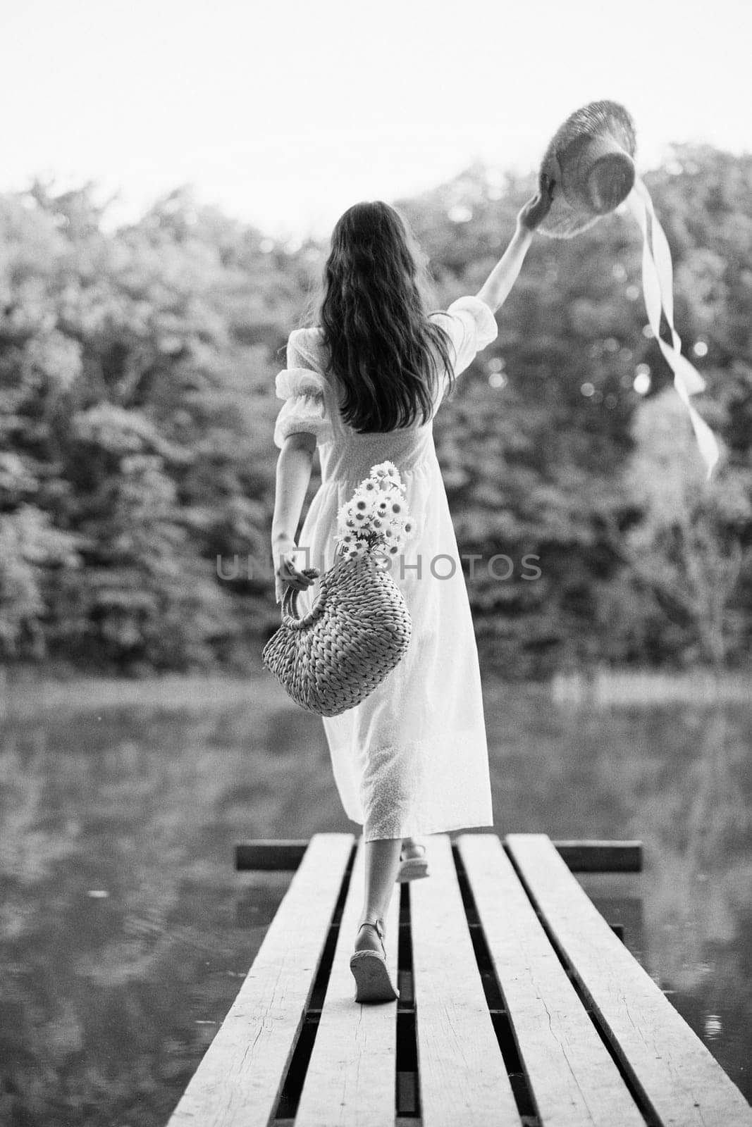 a woman in a long light dress stands on a pier by a lake in the forest with a basket and a wicker hat. monochrome photography by Vichizh