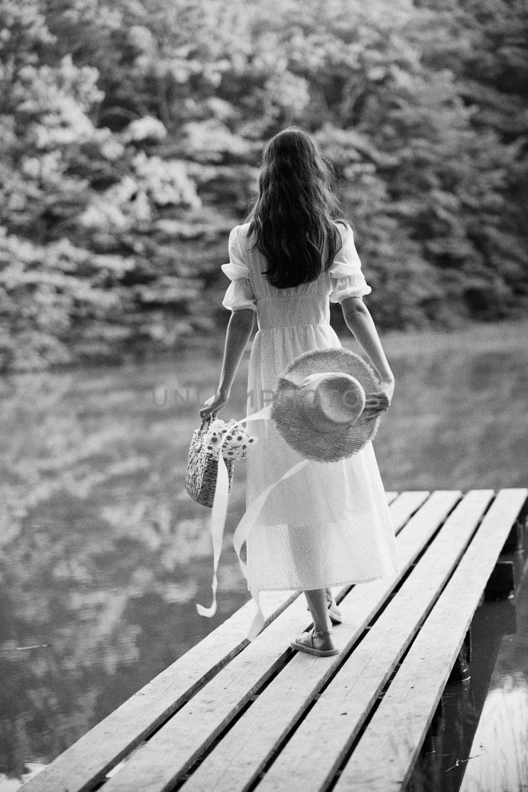 a woman in a long light dress stands on a pier by a lake in the forest with a basket and a wicker hat. monochrome photography. High quality photo