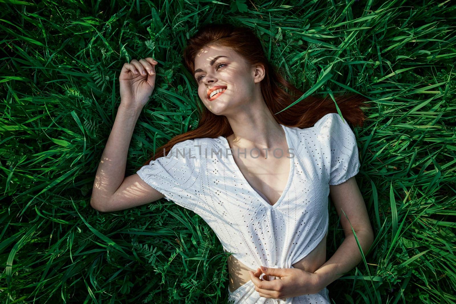 a beautiful woman with red hair lies on the grass in a white dress enjoying the day by Vichizh