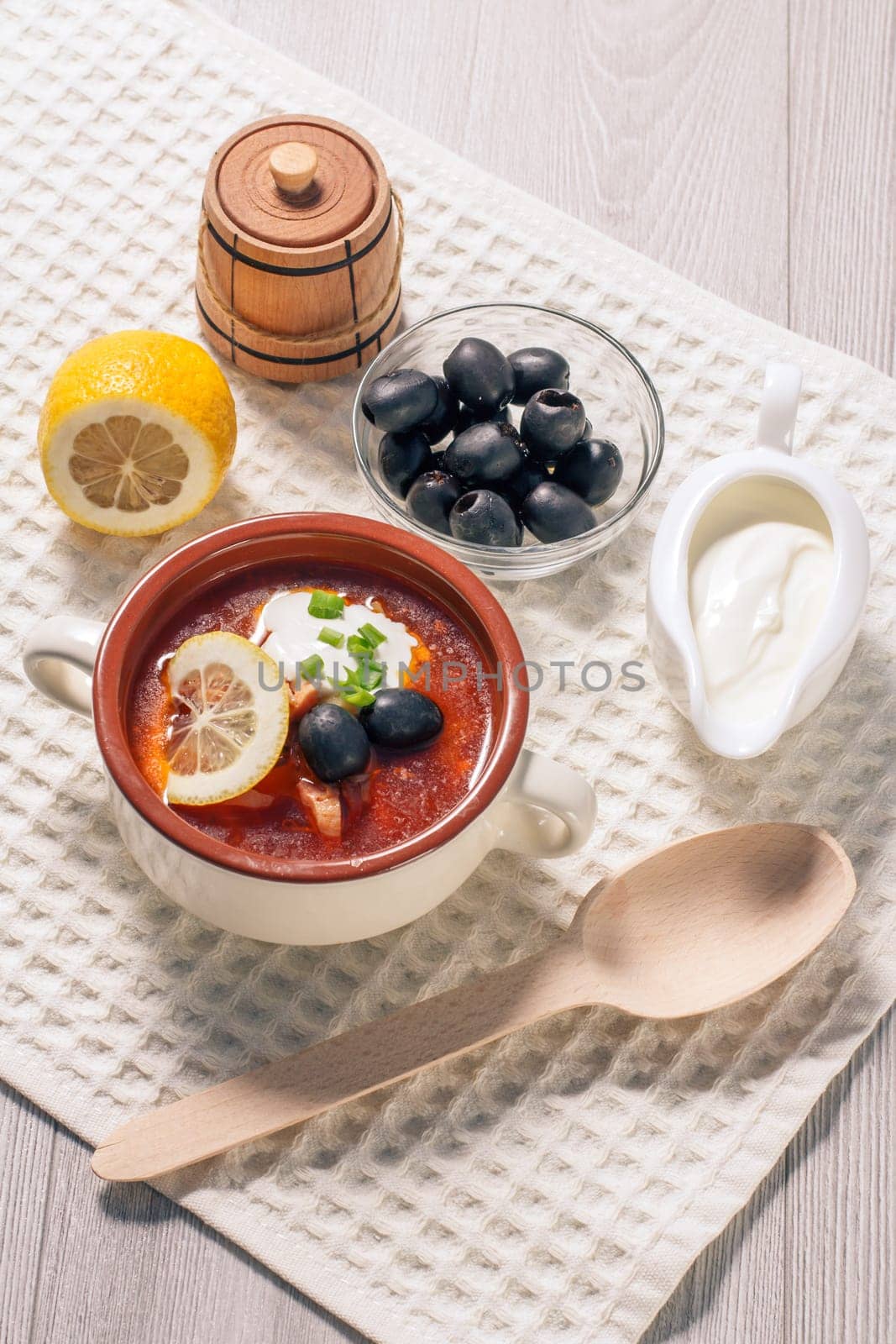 Soup saltwort with meat, smoked sausages, potatoes, tomatoes, marinated pickled cucumber, lemon, black olives and sour cream in ceramic soup bowl with ingredients on kitchen towel. Top view