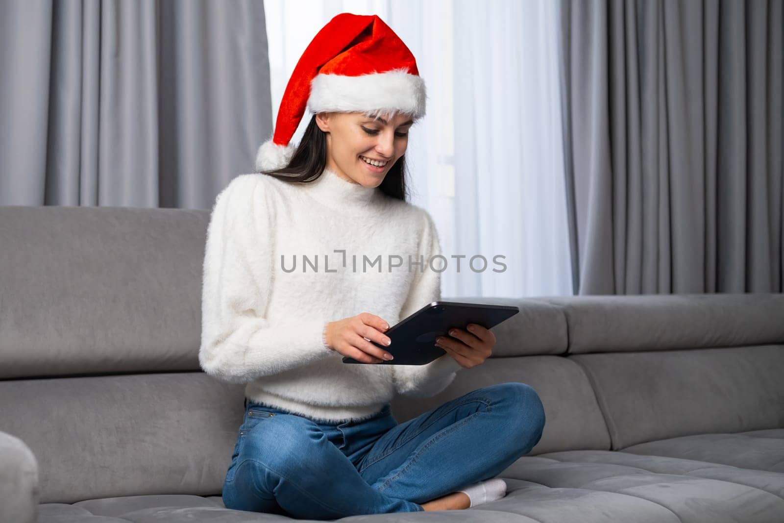 Happy young woman in a red Santa Claus hat sitting on the sofa and uses a tablet by vladimka