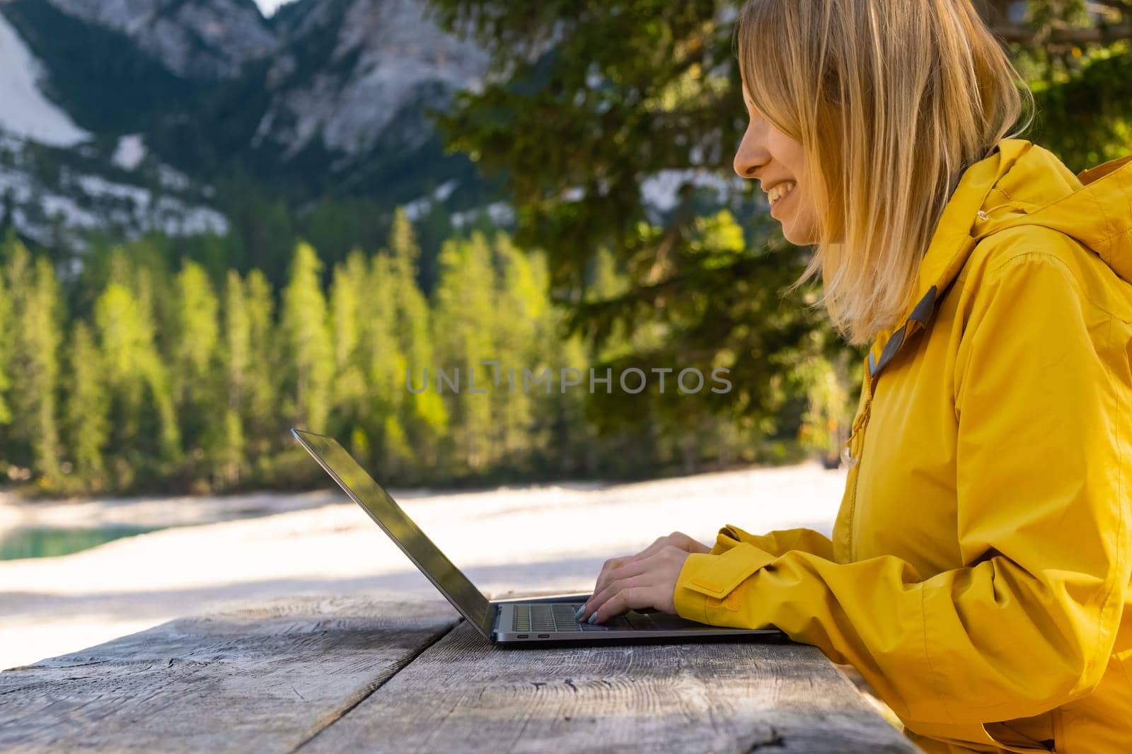 Smiling woman using her laptop with amazing view of mountains. Freelancer wearing yellow jacket and working remotely in the forest.