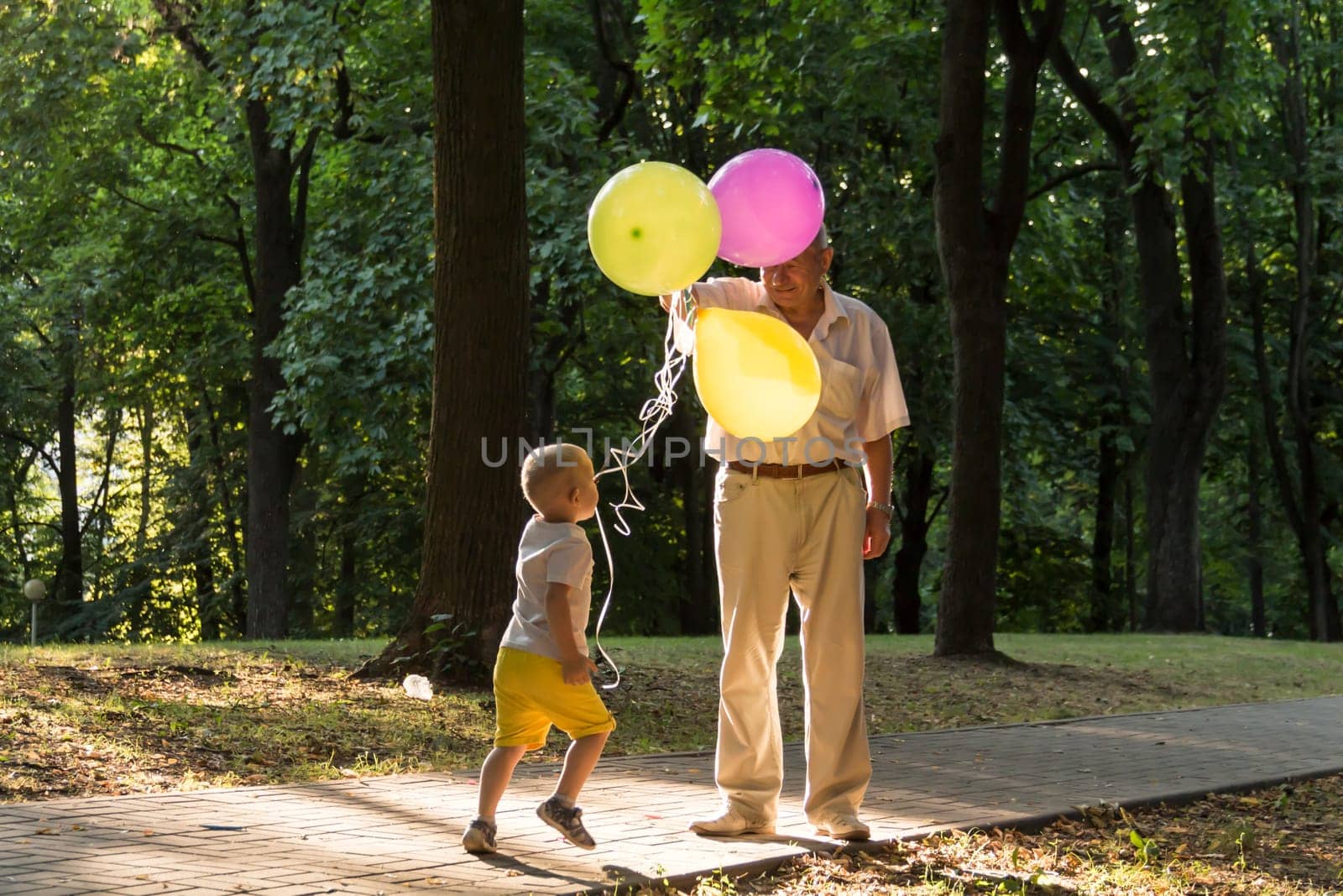 A little boy in yellow shorts and an elderly old man are playing with bright balloons. The family is having fun in the park in the fresh air and celebrating the holiday..