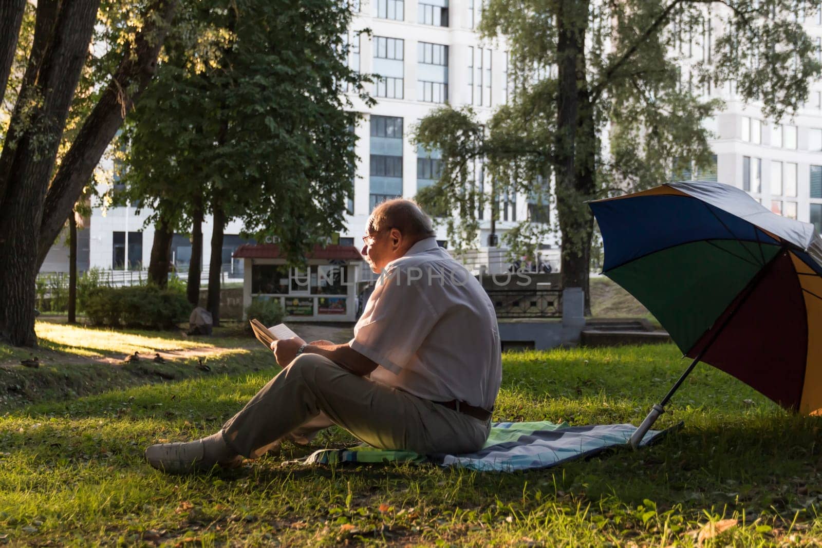 An elderly man in a white shirt is sitting on a blanket, on the ground in a park and reading an interesting book. A pensioner alone is resting in nature, passionate about his hobby.