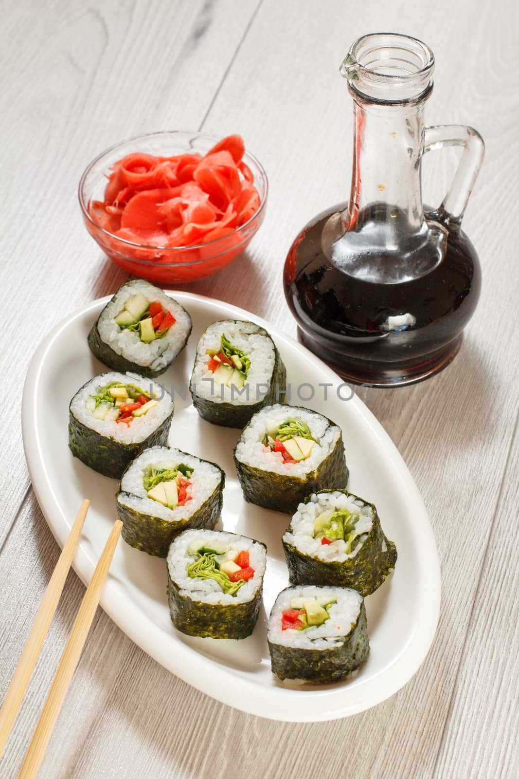 Sushi rolls with rice, pieces of avocado, cucumber, red bell pepper and lettuce leaves on ceramic plate, chopsticks, glass bottle with soy sauce and pickled ginger in a bowl. Vegetarian food