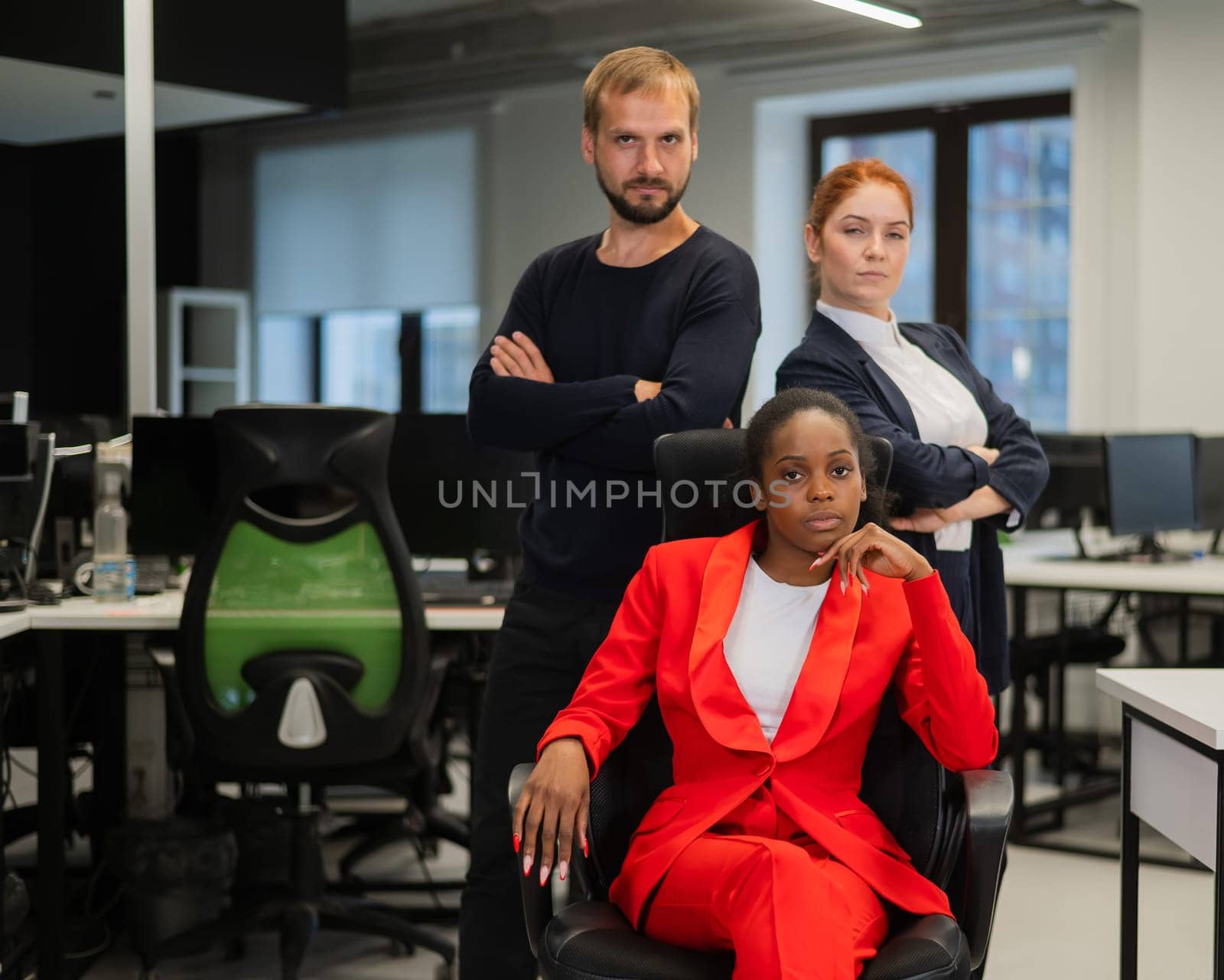 Caucasian red-haired woman, bearded Caucasian man stand behind a seated African American young woman in the office